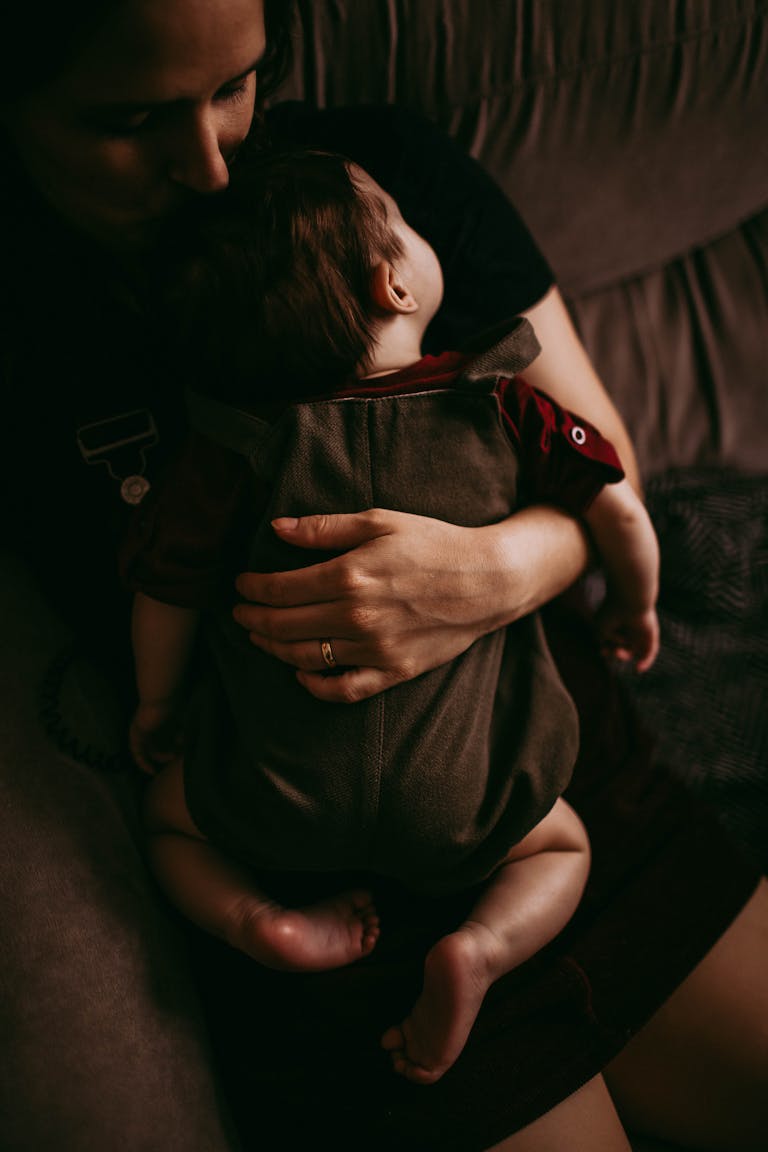 From above of crop unrecognizable mother kissing and cuddling napping baby on couch in apartment